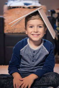 Portrait of smiling boy with book on head sitting at home