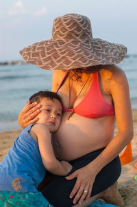 Son touching stomach of pregnant woman while sitting at beach against sky