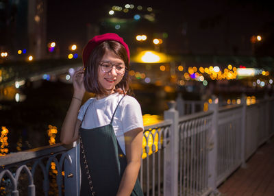 Thoughtful young woman smiling while standing against canal in illuminated city at night