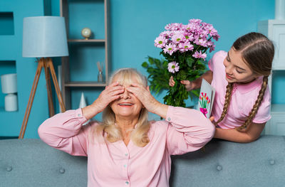 Cheerful grandmother closing eyes while granddaughter standing with flowers at home