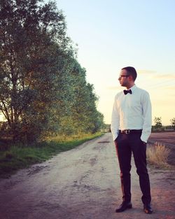 Full length of young man standing on dirt road by trees during sunset