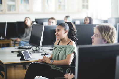 Smiling teenage girl sitting by young friend amidst computers at lab in high school