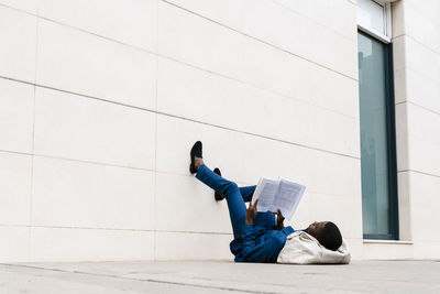 Male entrepreneur lying with duffel bag on sidewalk while reading book in city