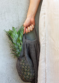 Female in linen white dress holds gray mash bag with ripe pineapple inside.