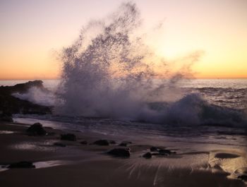 Waves splashing on shore against sky during sunset