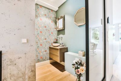 Doorway of stylish bathroom with bouquet of flowers and sink placed on cabinet at colorful wall with mirrors and wallpaper