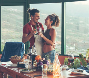 Smiling young couple toasting drinks while standing against window at home