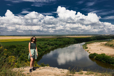 Portrait of woman standing at riverbank against cloudy sky