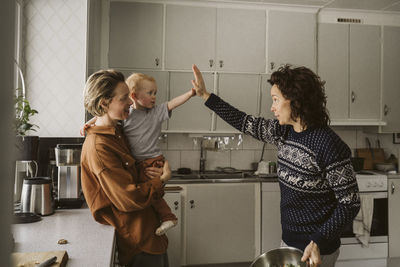 Woman carrying daughter giving high-five to mother in kitchen at home