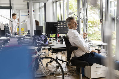 Businessman looking away while sitting on chair with colleagues in background at open plan office