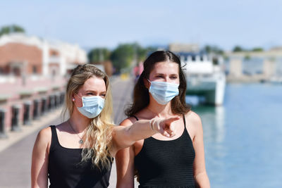 Portrait of young woman wearing mask pointing away while standing with friend at harbor