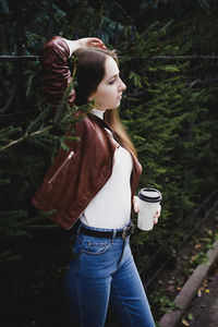 Beautiful young woman holding coffee cup while standing against plants