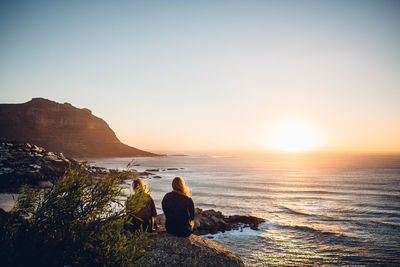 Rear view of people sitting on rock by sea against sky during sunset