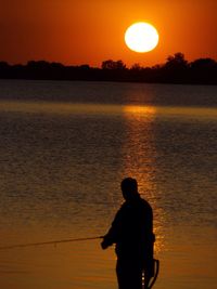 Silhouette fisherman standing against lake during sunset