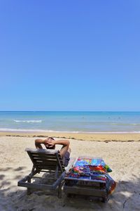 Man relaxing on lounge chair at beach against clear blue sky