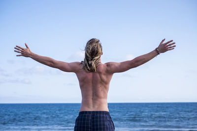 Rear view of shirtless man standing with arms outstretched against sky