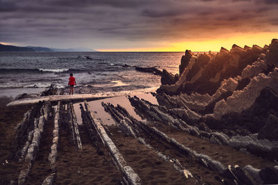 Rear view of boy standing at beach against cloudy sky during sunset