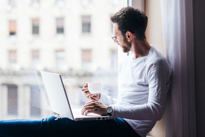 Man using laptop while sitting on window sill