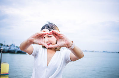 Portrait of woman making heart shape with hands while standing against sea