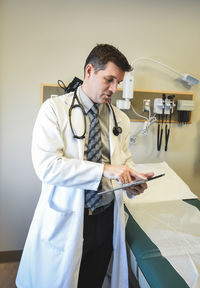 Doctor in white coat beside exam table in hospital looking a tablet.