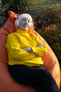 Attractive gray-haired 85 year old woman sits on a bag chair and listens to music on headphones.