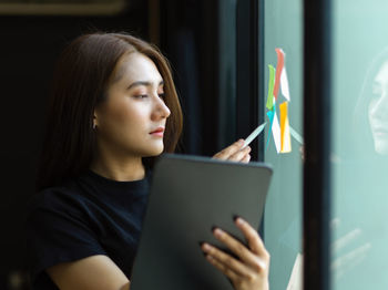 Young woman holding digital tablet while writing over sticky notes on window