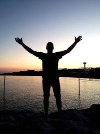 Silhouette of man standing with arms outstretched on shore against sky during sunset