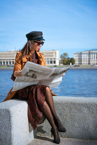 Stylish young woman reading fresh newspaper on embankment of river on sunny day