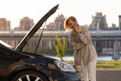 Woman talking over phone by breakdown car on road in city