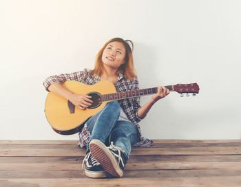 Smiling young woman playing guitar while sitting against white wall