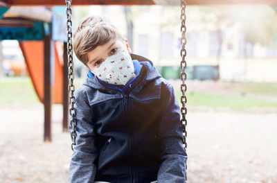 Boy wearing face mask and feeling lonely while sitting on a swing at the park during coronavirus.