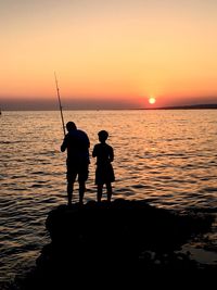 Silhouette man with son fishing at beach against sky during sunset