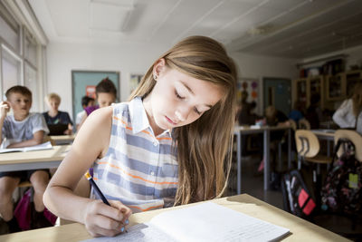 Girl writing in book at desk with students sitting in background