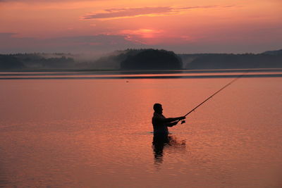Silhouette man fishing in lake against orange sky