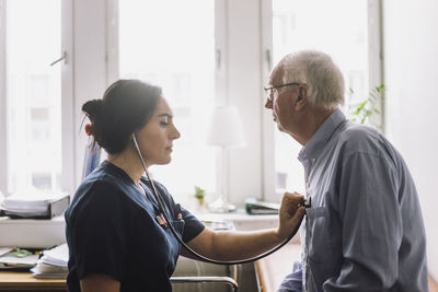 Side view of female nurse listening heartbeat of senior male patient in clinic