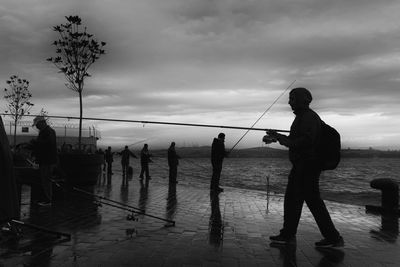 Silhouette people fishing in river on pier against cloudy sky