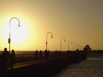 Silhouette people on street by sea against clear sky during sunset