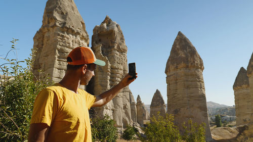 A man in a yellow t-shirt and baseball cap takes a picture of mountains with his phone. love valley