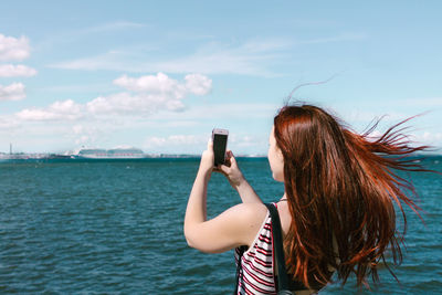 Woman photographing with mobile phone by sea against sky