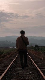 Rear view of man walking on railroad track against sky during sunset