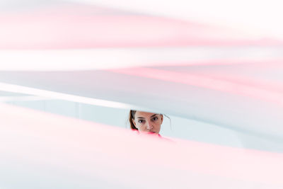 Crop attractive female looking at camera with serious face through translucent light textile while standing near white wall in light studio