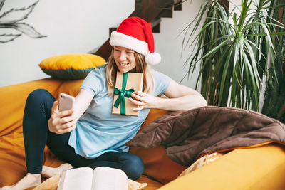 A smiling girl in a christmas hat sits on the sofa and communicates via video link with friends 