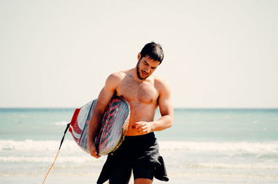 Young man with surfboard standing on beach against sky