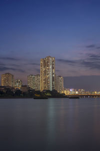 Illuminated modern buildings by bay against sky at dusk