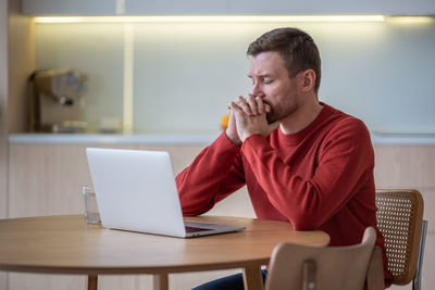 Sad worried man sits with laptop at table with eyes closed thinking of project failure, bad luck.