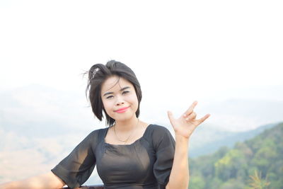 Portrait of smiling young woman gesturing horn sign while standing against mountain and sky