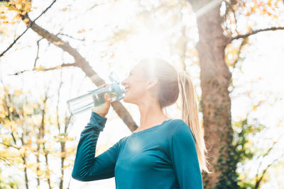 Young woman drinking water while standing against trees