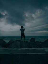 Rear view of man standing on beach against cloudy sky