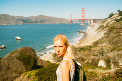 Portrait of woman standing at sea shore by golden gate bridge against clear sky
