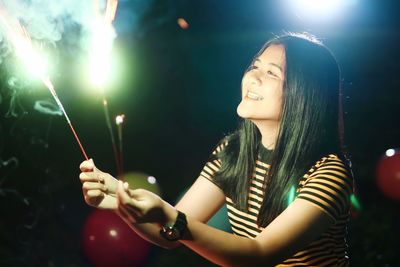 Smiling young woman burning sparklers while standing outdoors at night
